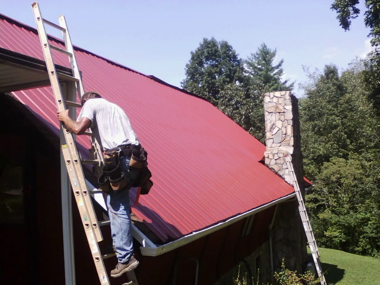 Roofer on a ladder beside a red metal roof and stone chimney — Soffit & Fascia Repair by Roofing Brothers in Lulu